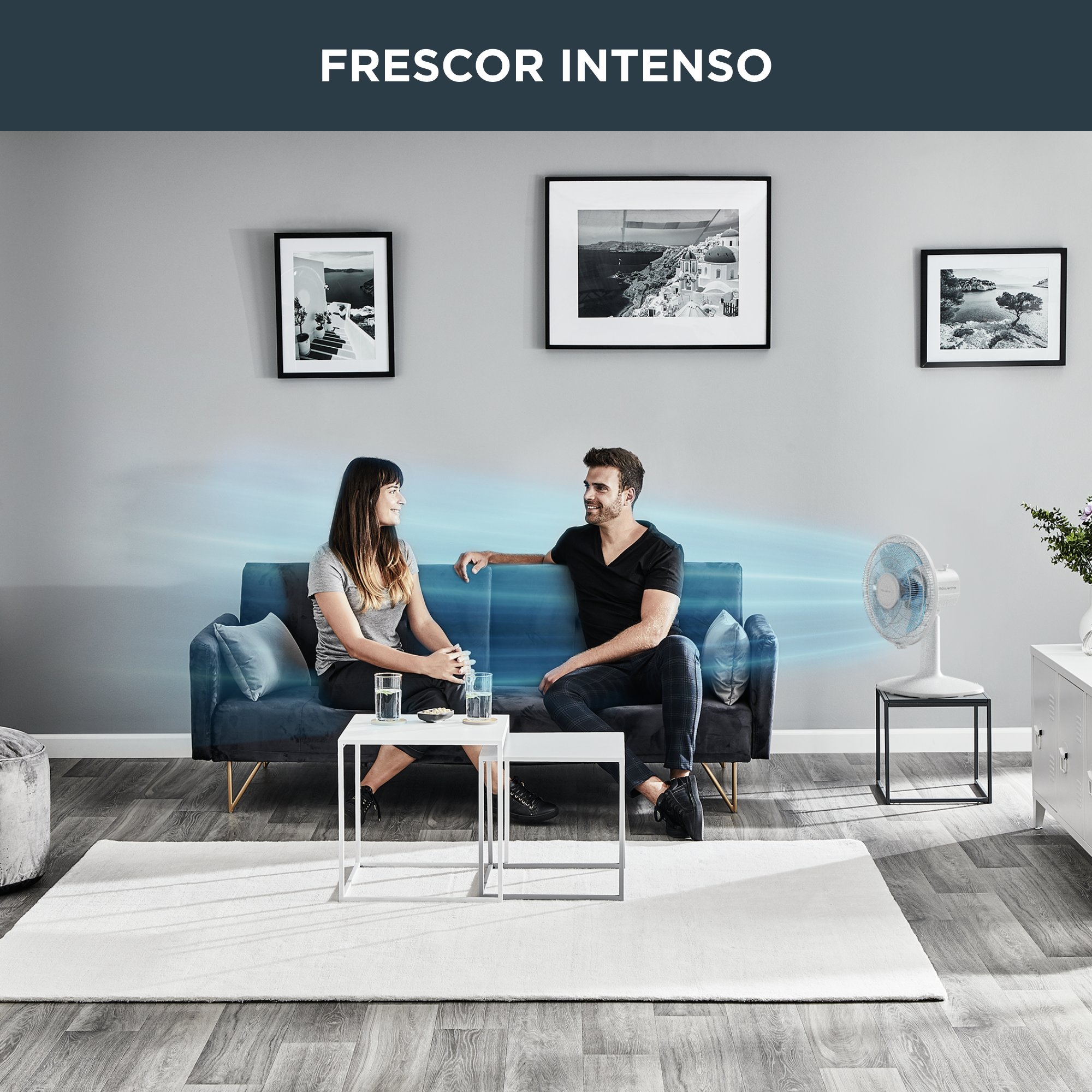 Dos personas sentadas en un sofá azul frente a un ventilador. Cuadros en la pared.