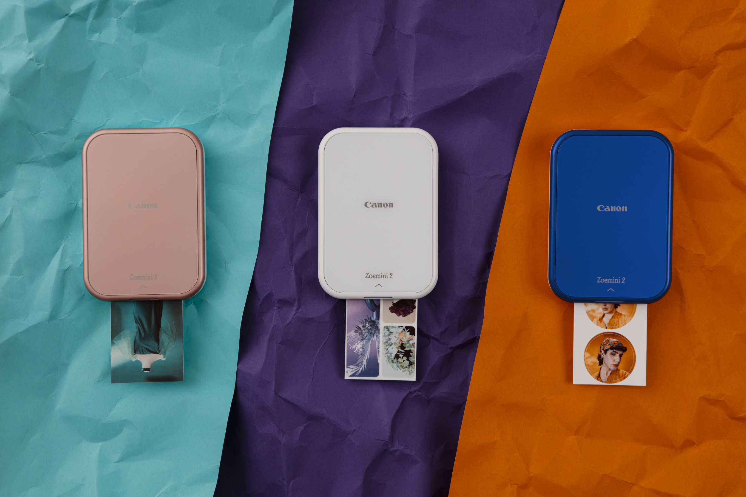 Drei Canon Zoemini 2 Drucker, verschiedene Farben, auf buntem Papier.