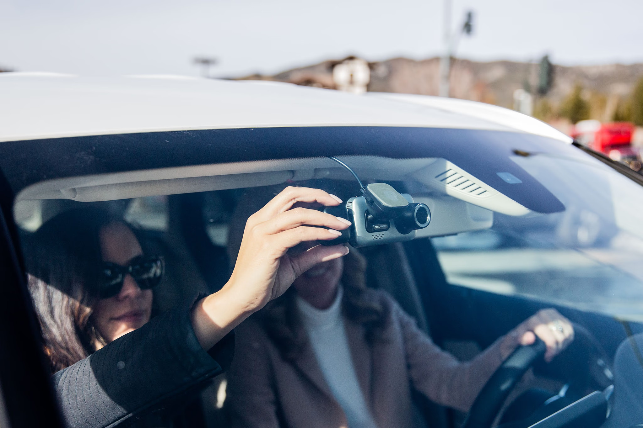 Deux femmes dans une voiture installant une caméra embarquée sur le pare-brise. Extérieur, journée ensoleillée.
