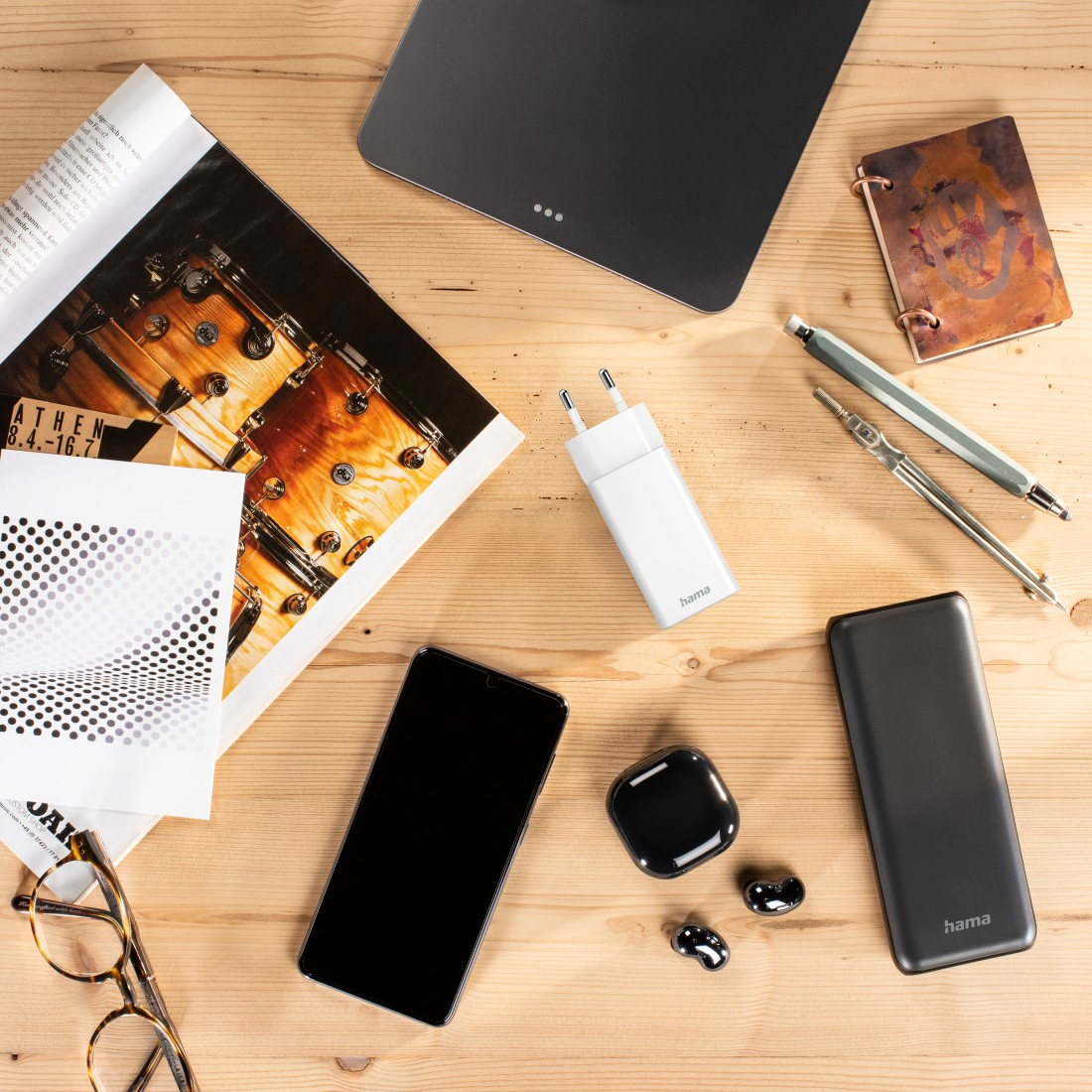 Overhead shot of accessories: phone, charger, earbuds, pens, notepad, magazine on a wooden table.