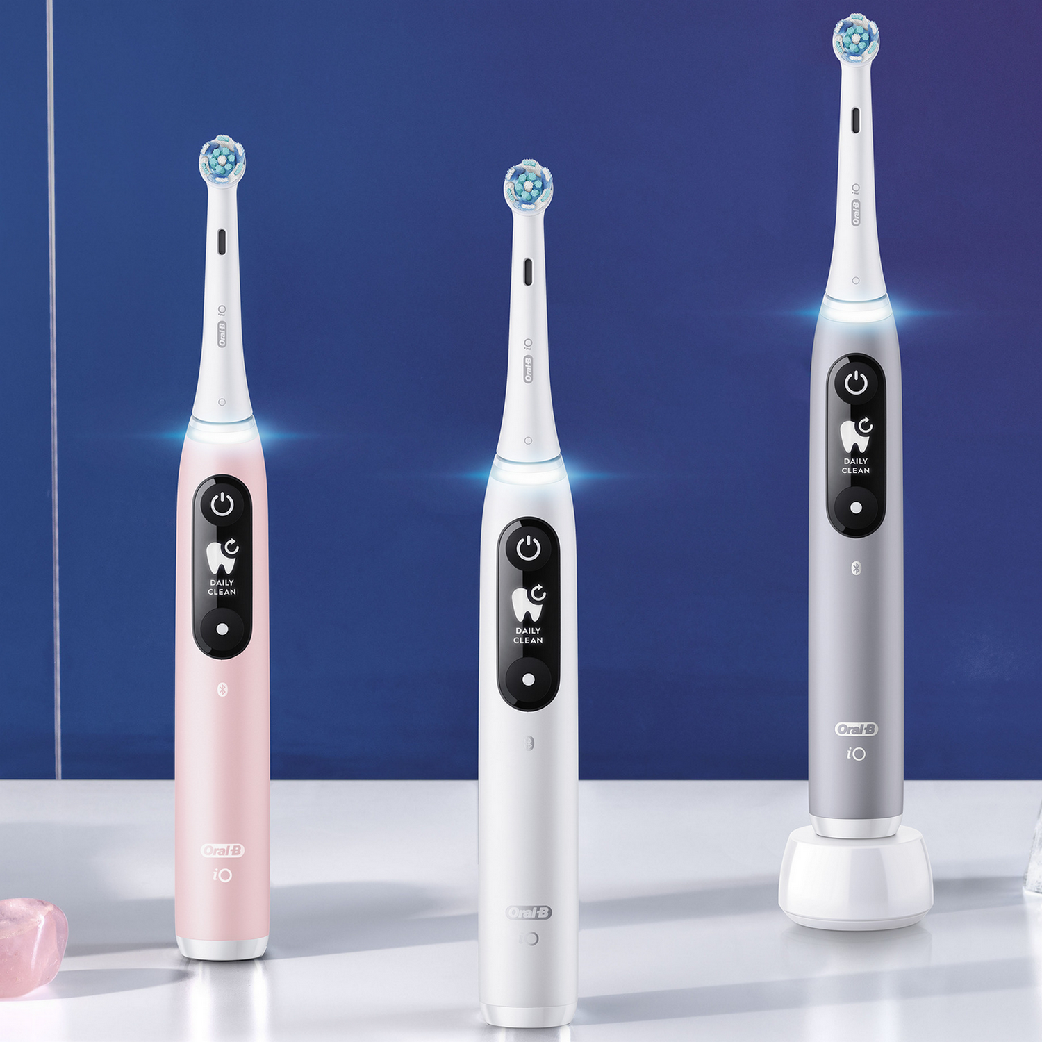 Three electric toothbrushes, pink, white, and gray, with a charging base. All are on a table.