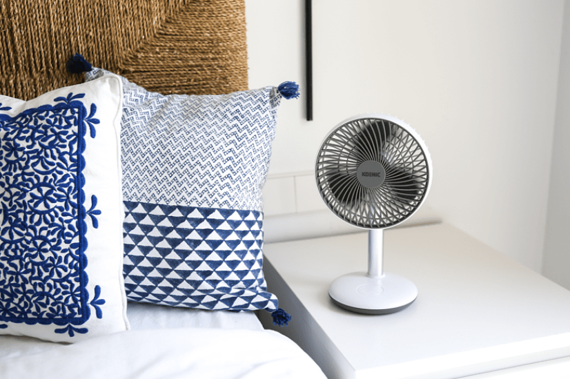 Fehér ventilátor egy fehér éjjeliszekrényen, párnák mellett az ágyon. Szobai beállítás.