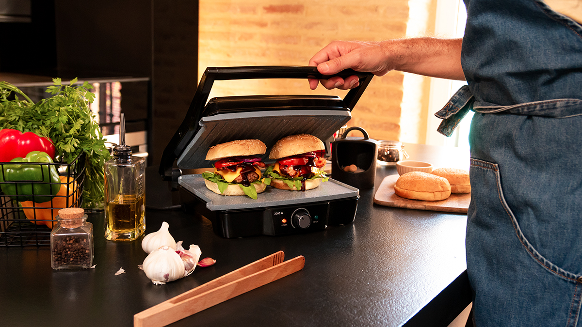 Persona asando hamburguesas en una plancha. Ingredientes y utensilios están en la mesa.