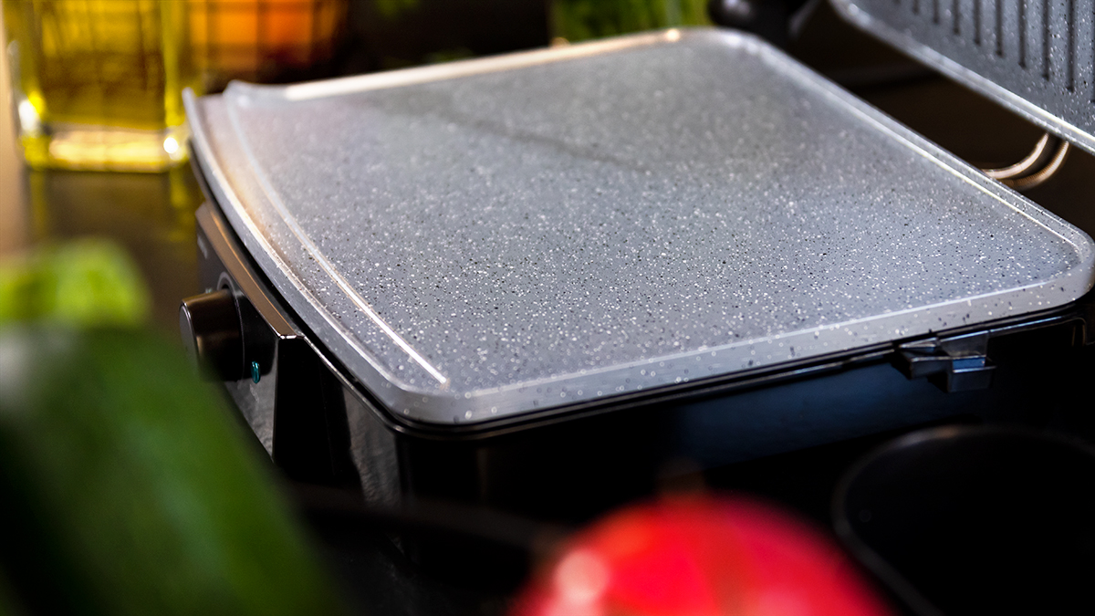 Primer plano de una placa de parrilla gris con un patrón moteado. En el fondo, se ven alimentos y utensilios de cocina.