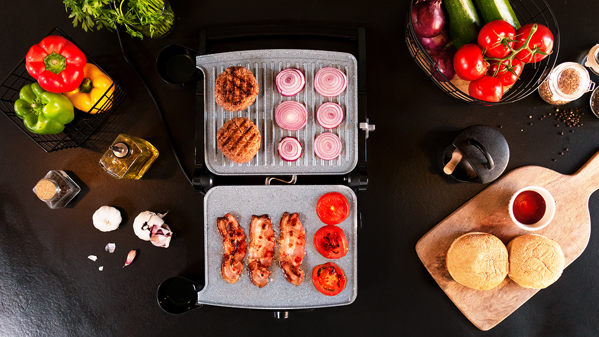 Vista cenital de la parrilla. Se están asando hamburguesas y cebollas, tomates y tocino. Panecillos y condimentos.