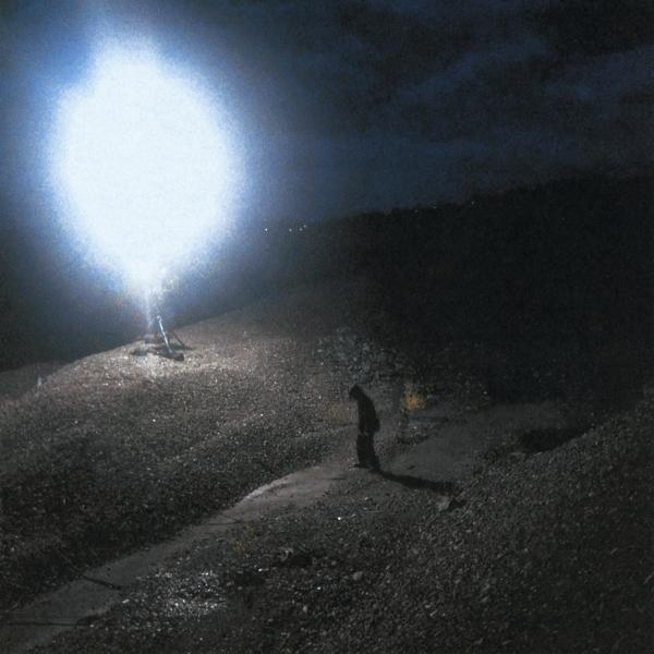 Dunkle Nachtszene. Eine einsame Gestalt steht auf einem Weg, beleuchtet von einer hellen, runden Lichtquelle.