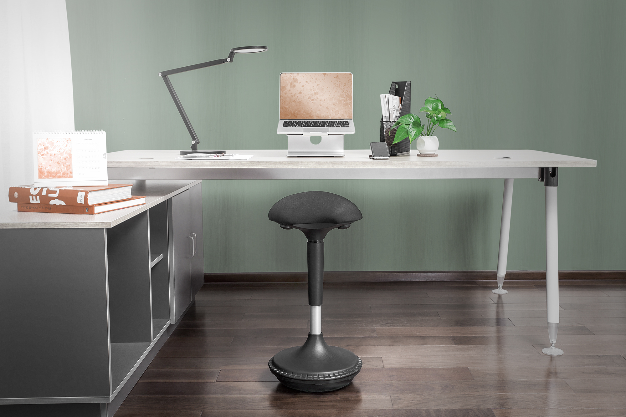 Bürotisch mit Laptop, Lampe und Pflanzen. Ein schwarzer Hocker steht davor. Grüne Wand im Hintergrund.