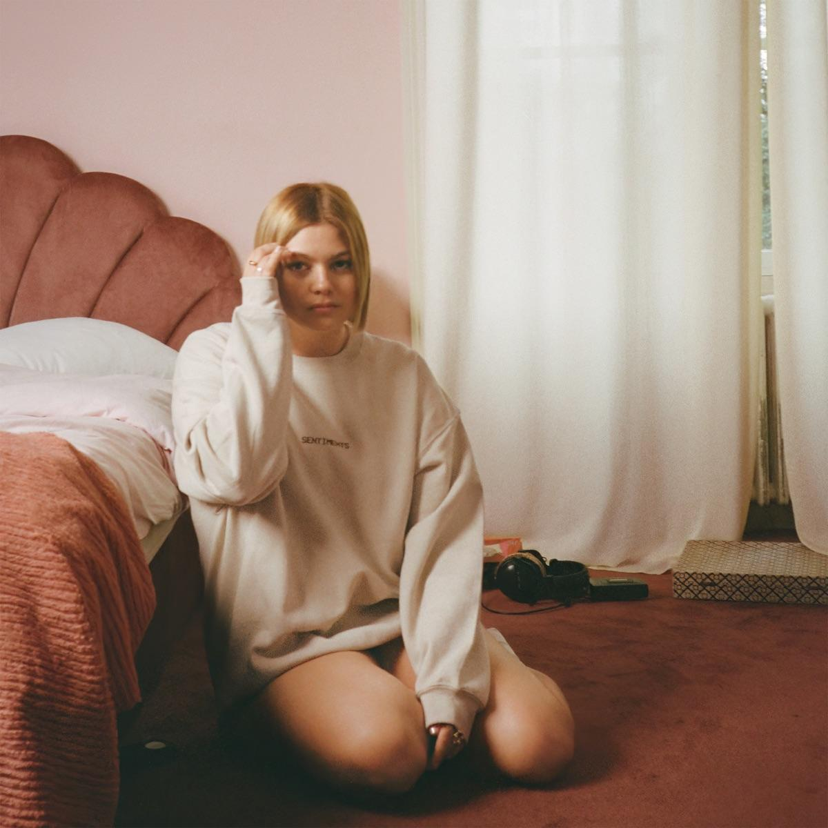 Femme agenouillée sur le sol, portant un sweat-shirt, dans une chambre rose. Cadre de chambre.