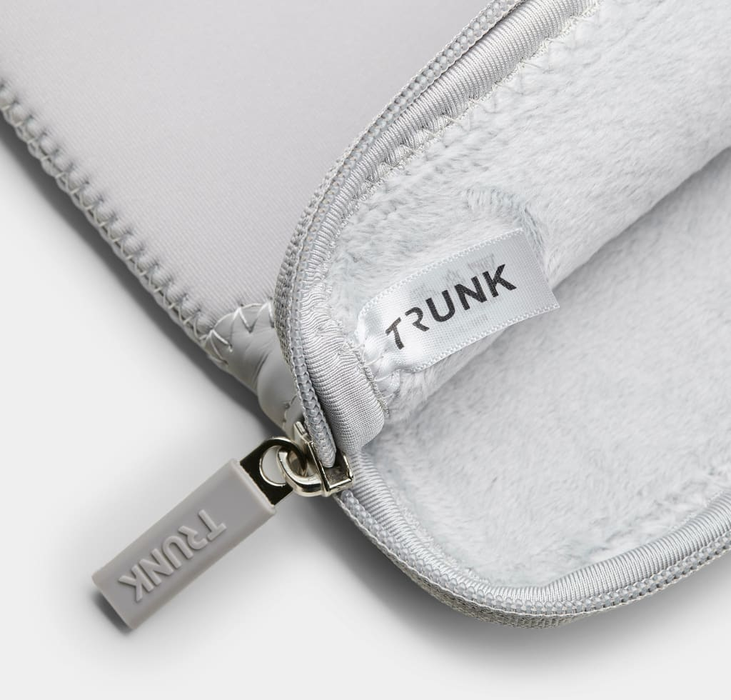 Eine hellgraue Laptoptasche ist geöffnet und zeigt ein weiches, weißes Innere. Das Wort TRUNK steht auf einem Etikett.