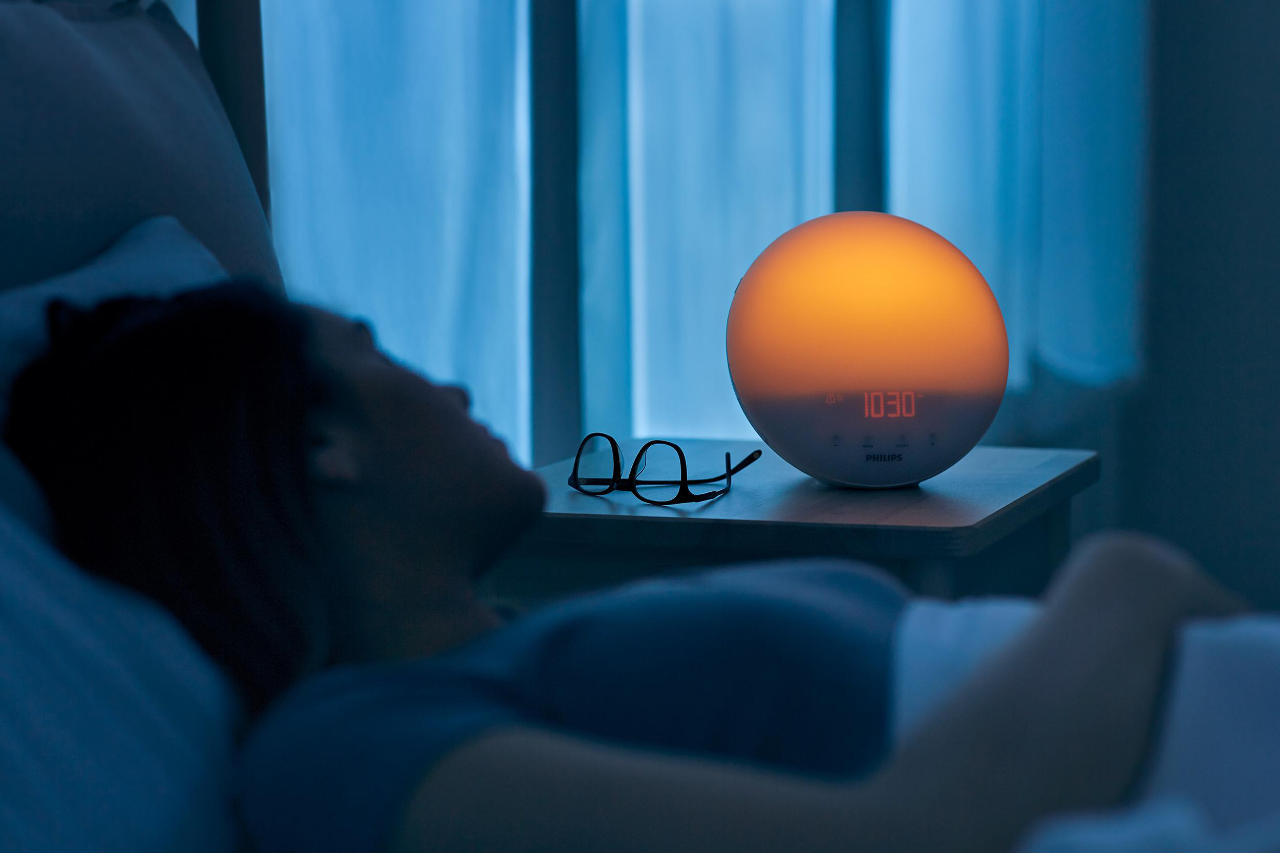Eine Person schläft in einem Bett, während ein Sonnenaufgang-Wecker auf einem Nachttisch steht.
