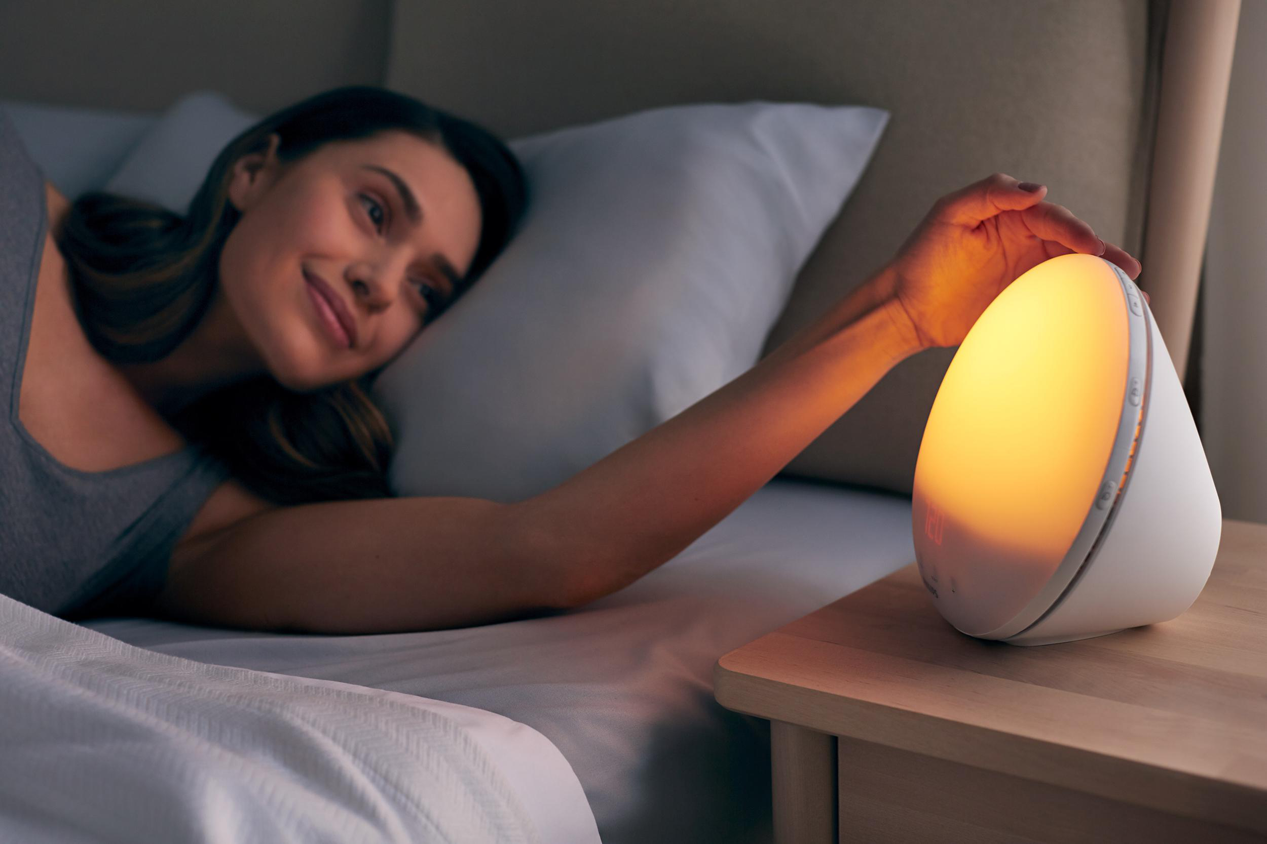 Eine Frau im Bett greift nach einer weißen Nachttischlampe. Die Lampe strahlt warmes, orangefarbenes Licht aus.