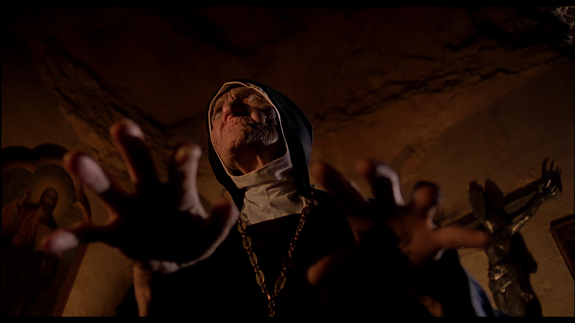 Eine Nonne in einem dunklen Raum, die mit Händen auf den Betrachter zugeht, im Hintergrund ein Kruzifix.