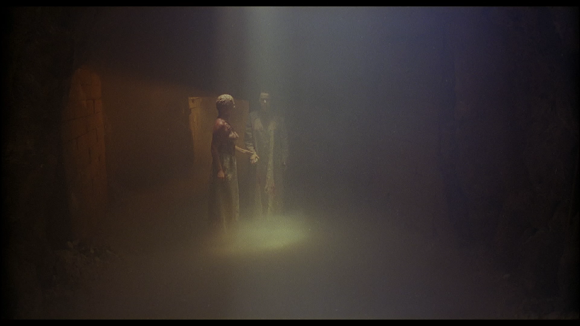 Zwei Figuren in einem schwach beleuchteten Raum, umgeben von Nebel. Licht scheint vom Boden.