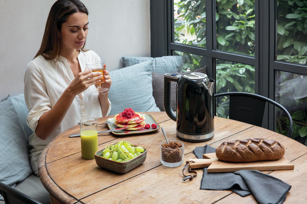 Frau am Holztisch mit Pfannkuchen, Saft und Brot, trinkt Tee neben einem schwarzen Wasserkocher.