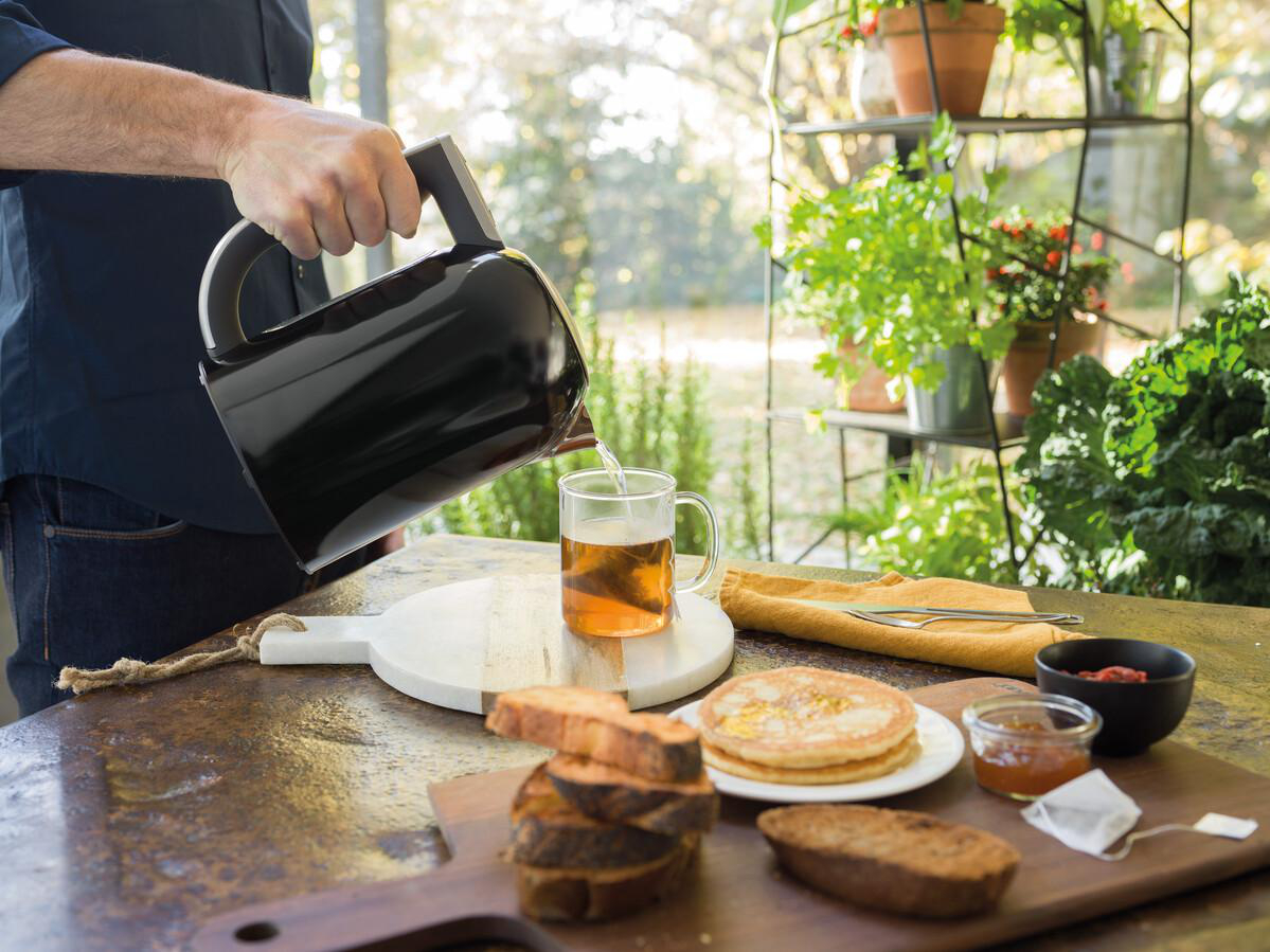 Person gießt Tee in eine Tasse. Ein Frühstücksarrangement mit Toast, Pfannkuchen und Marmelade wird gezeigt.