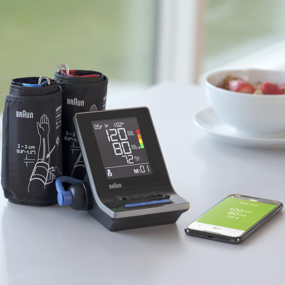 Ein Blutdruckmessgerät mit zwei Manschetten und einem Smartphone, das Messwerte anzeigt. Im Hintergrund eine Schüssel mit Essen.
