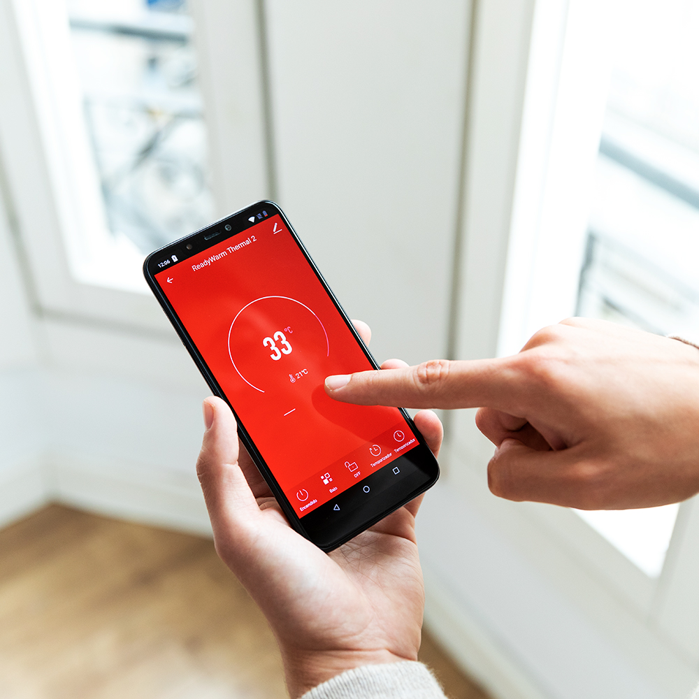 Una mano sostiene un teléfono mostrando 33°C en una pantalla roja. Otra mano toca la pantalla. El fondo es una ventana.