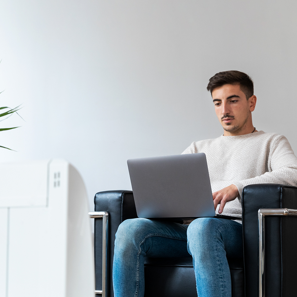 Hombre con suéter blanco y jeans sentado en una silla negra, trabajando en una laptop. Un muro blanco en el fondo.