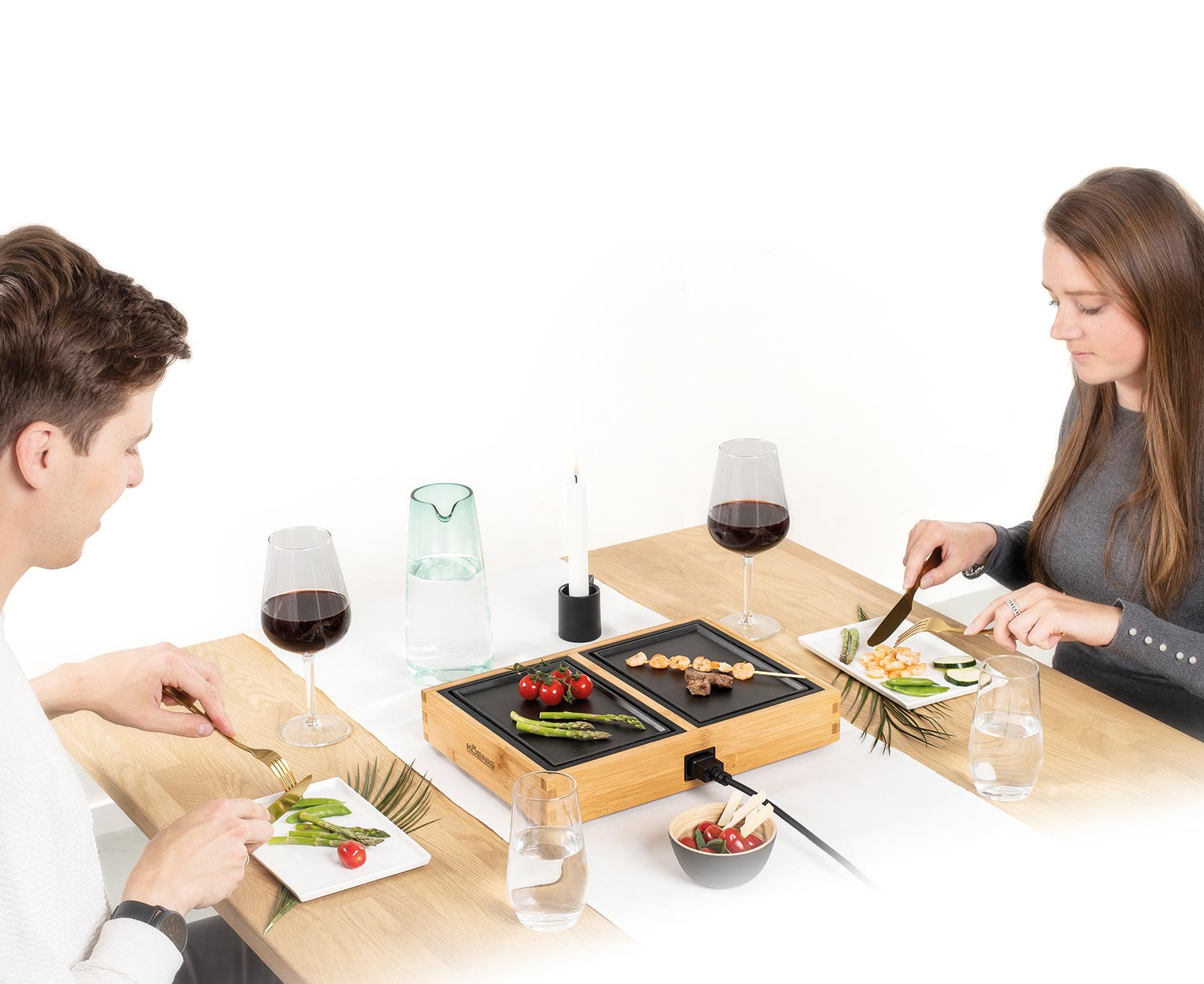 Un couple mangeant à une table avec un petit grill. Verres à vin et nourriture sur la table. Fond blanc.