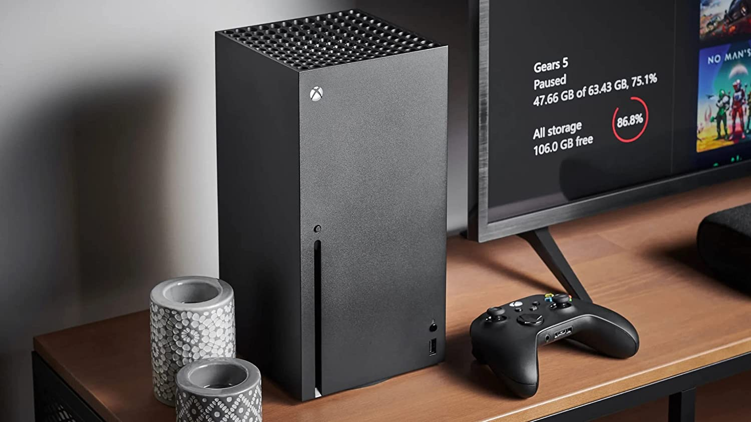 Schwarze Xbox Series X Konsole. Controller auf Holztisch, TV im Hintergrund mit Spielbildschirm.