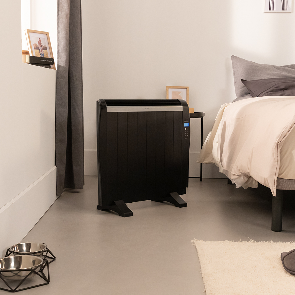 Un calefactor negro en un dormitorio. Cama con cabecero gris y manta blanca a la derecha. Comederos para perros delante.