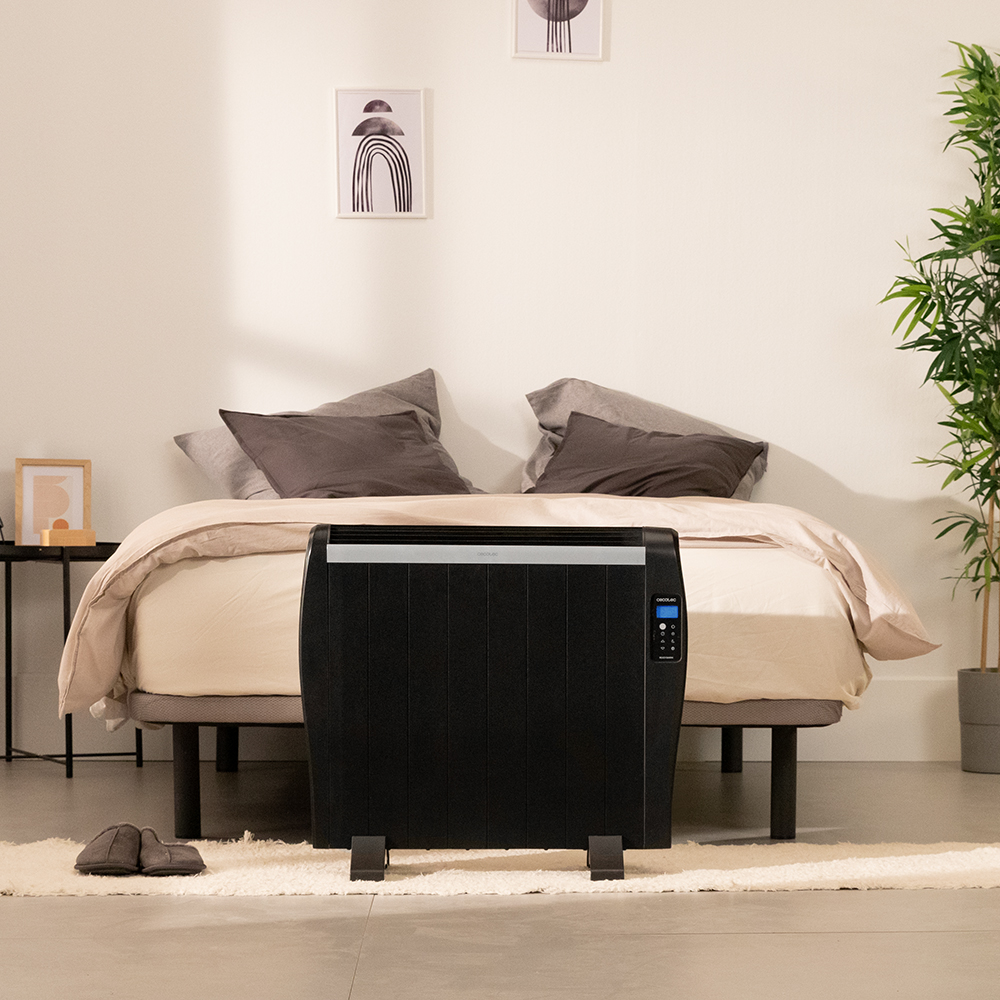 Un calefactor negro frente a una cama con almohadas grises y una manta beige. También se ven una planta y arte.