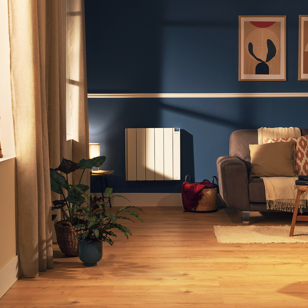 Salón con pared azul, plantas, sofá, calefactor y obras de arte. Suelo de madera, cortinas beige y luz cálida crean un ambiente acogedor.