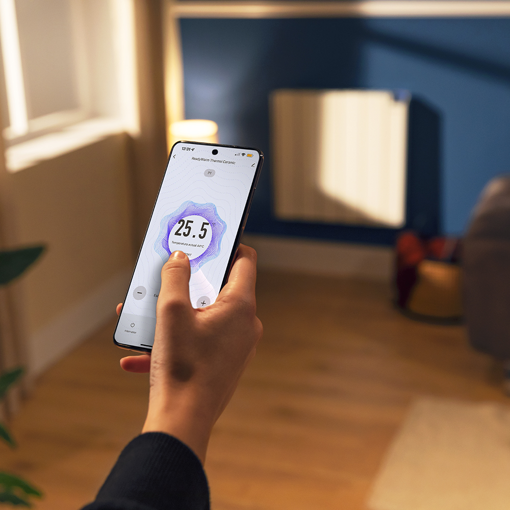 Una persona sostiene un teléfono que muestra una lectura de temperatura. En el fondo, un calefactor en una habitación pintada de azul.