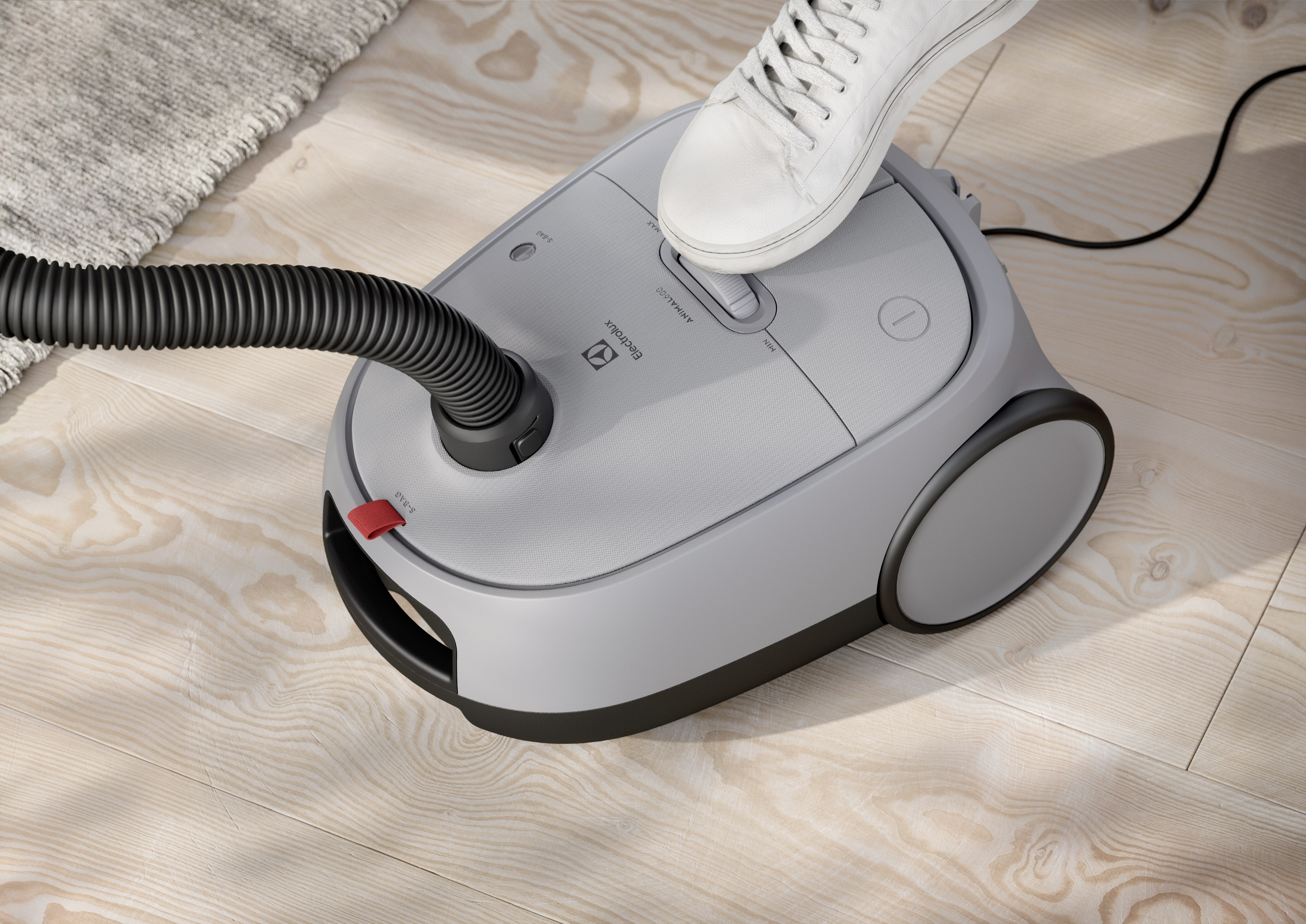Pied sur un aspirateur gris, tuyau attaché, sur plancher en bois.