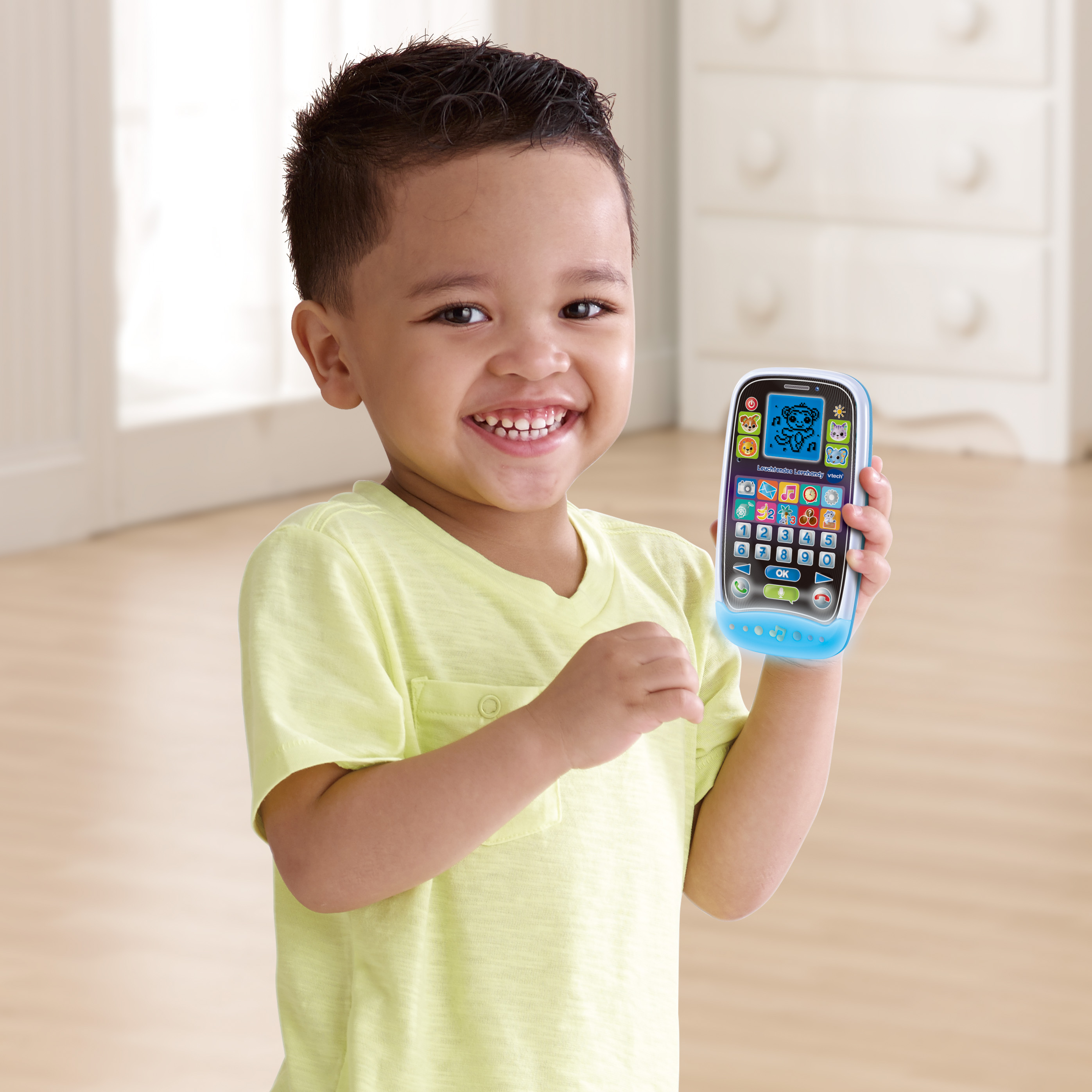 Ein kleiner Junge lächelt und hält ein blaues Spielzeugtelefon. Er trägt ein gelbes Shirt.