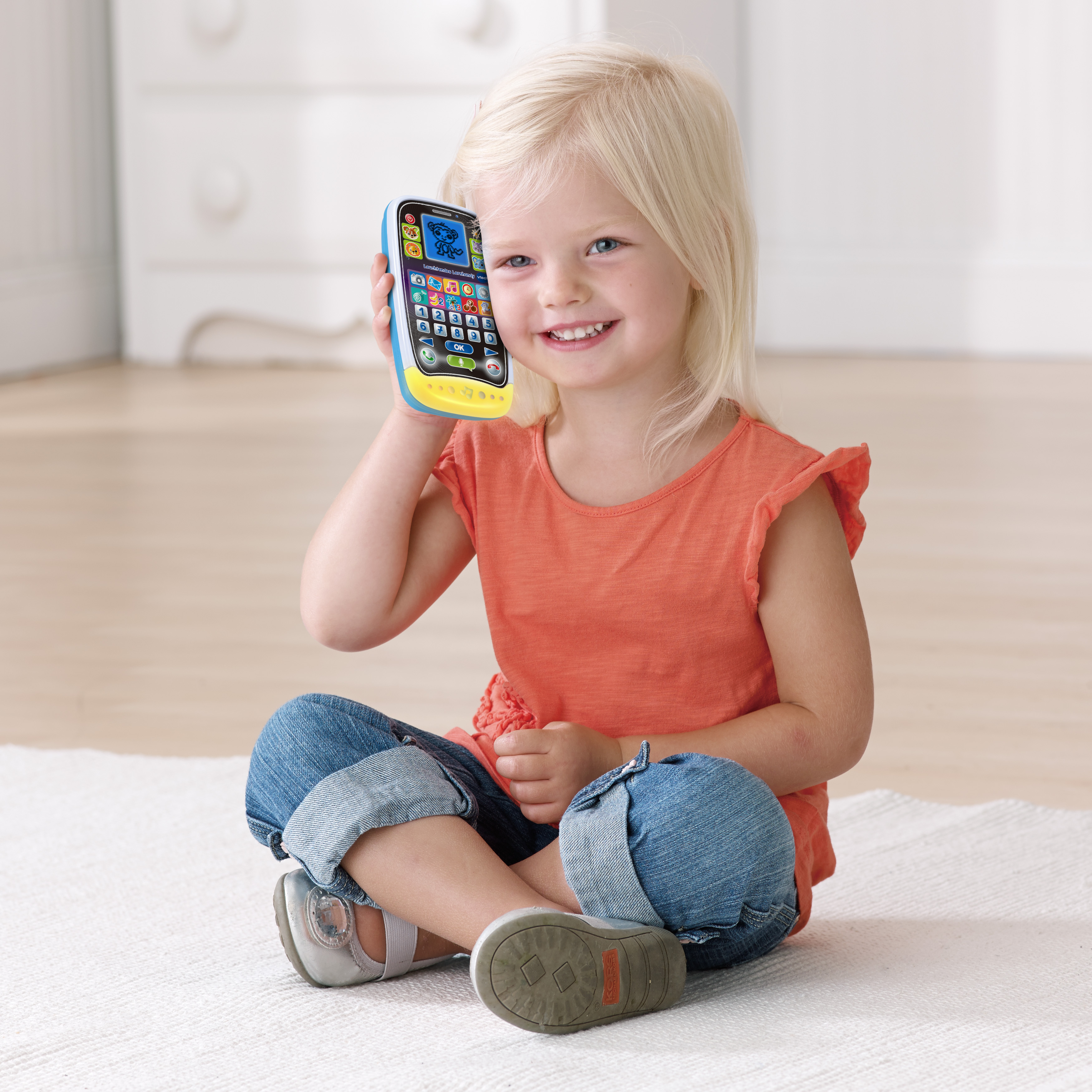 Blondes Mädchen hält ein Spielzeugtelefon. Sitzt auf einem weißen Teppich.