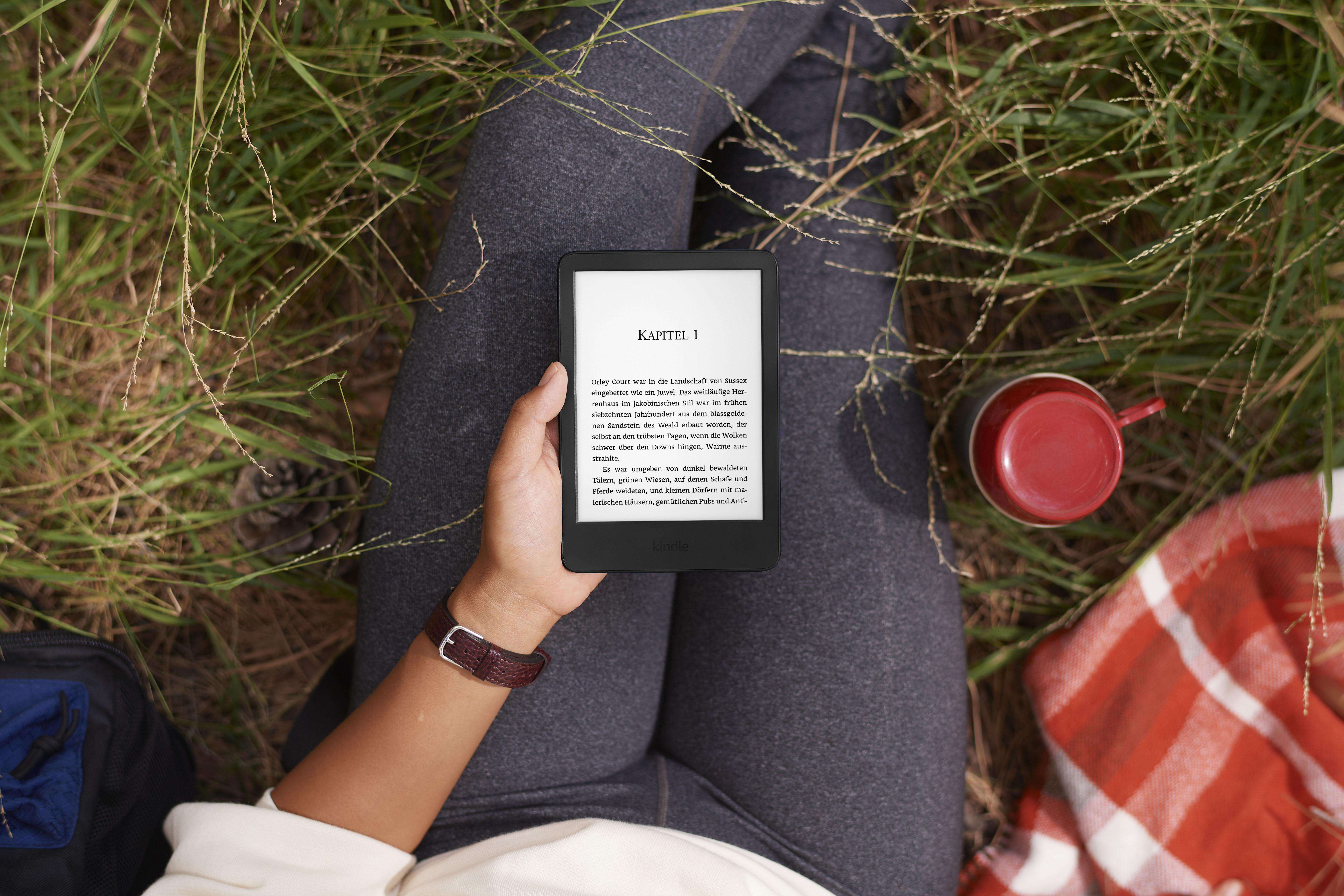 Eine Person liest ein Buch auf einem Kindle-Gerät, während sie mit einem roten Becher im Gras liegt.
