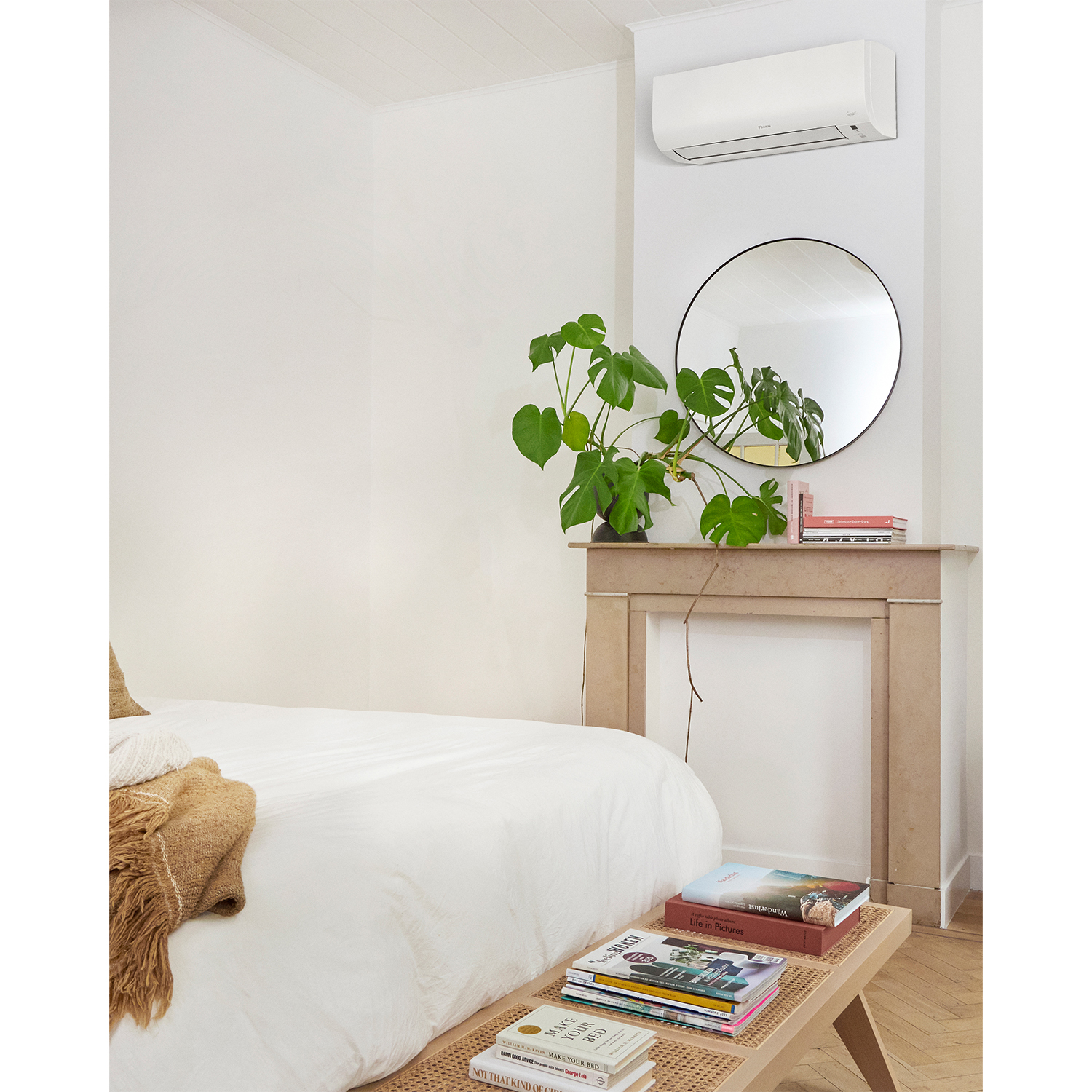 Camera da letto con letto, pianta, specchio e libri su una panca. Condizionatore sopra lo specchio.