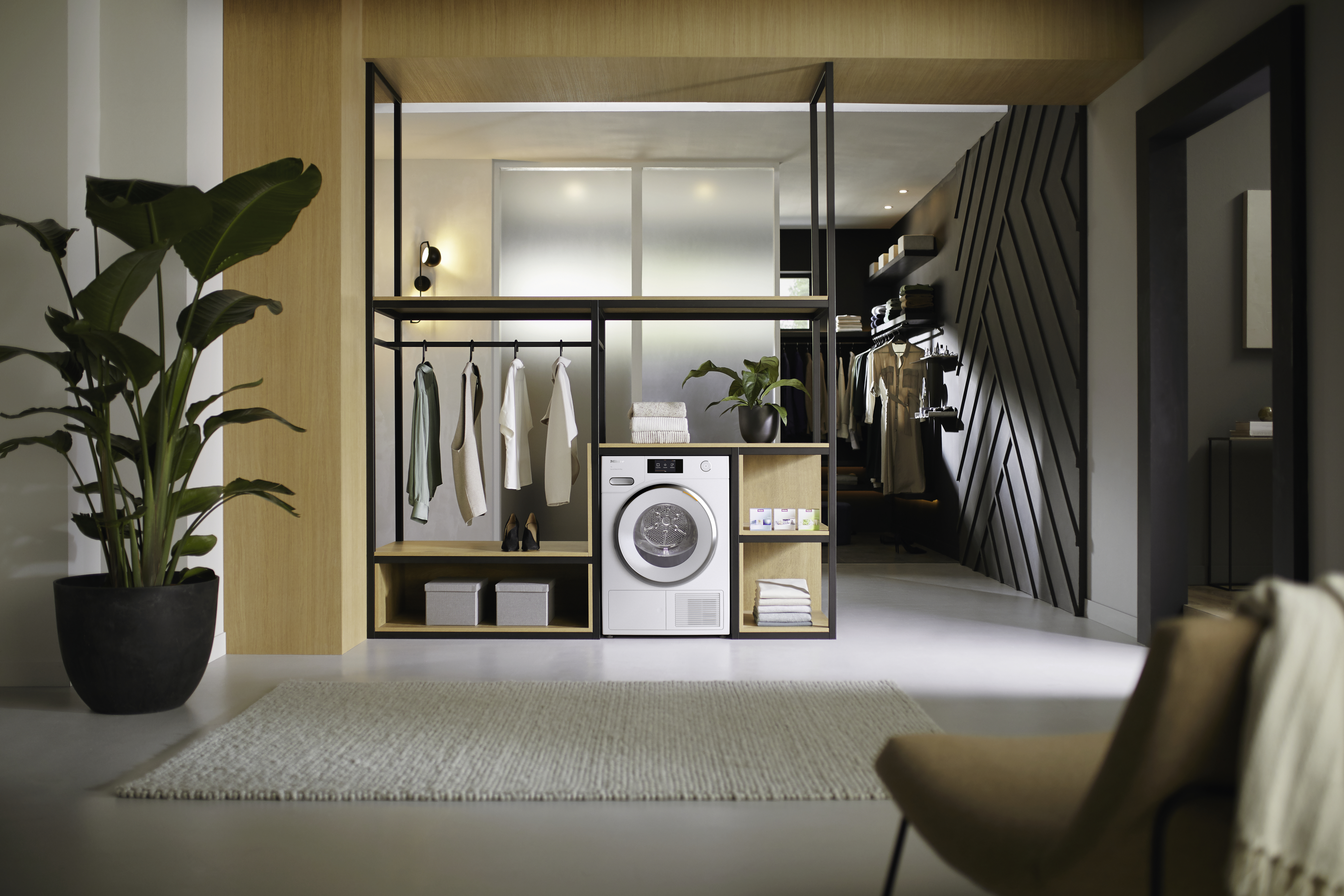 Modernes Interieur mit Waschmaschine, Kleidung auf einem Gestell und einem lichtdurchfluteten Wohnraum.