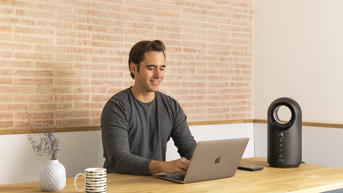 Un hombre sonríe mientras usa una laptop en un escritorio de madera. Un ventilador negro está a la derecha. Una pared de ladrillos está en el fondo.