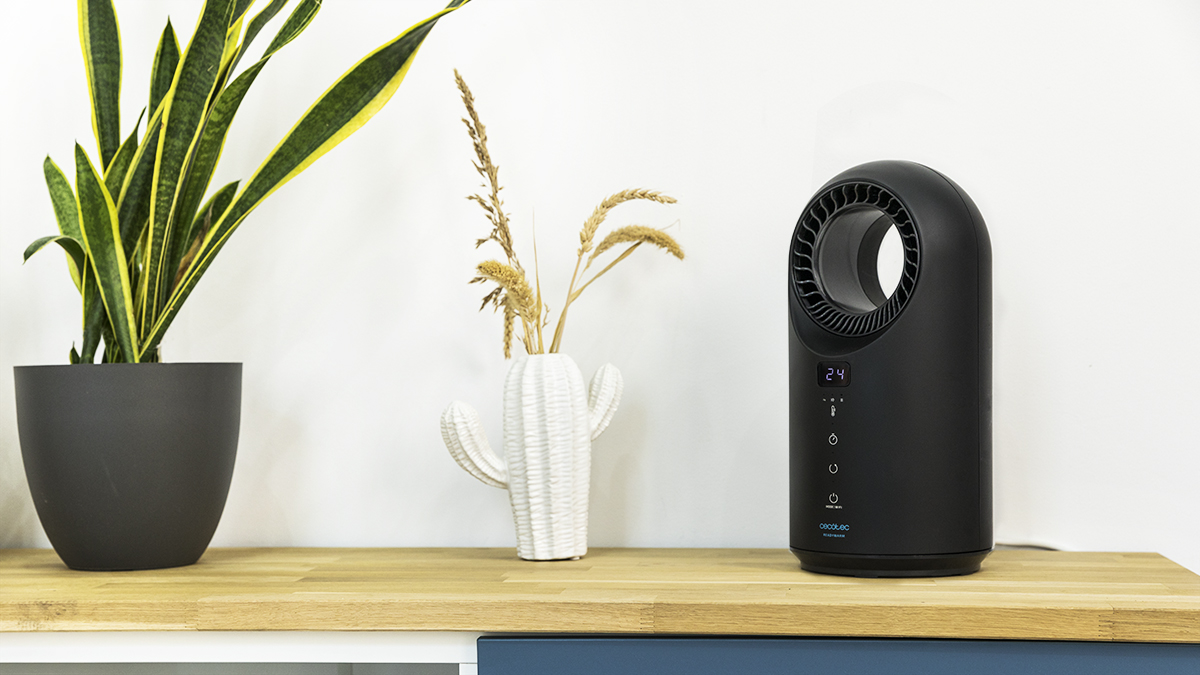 Un ventilador negro con pantalla digital está sobre una superficie de madera. Un jarrón en forma de cactus con flores secas y una planta están a la izquierda.