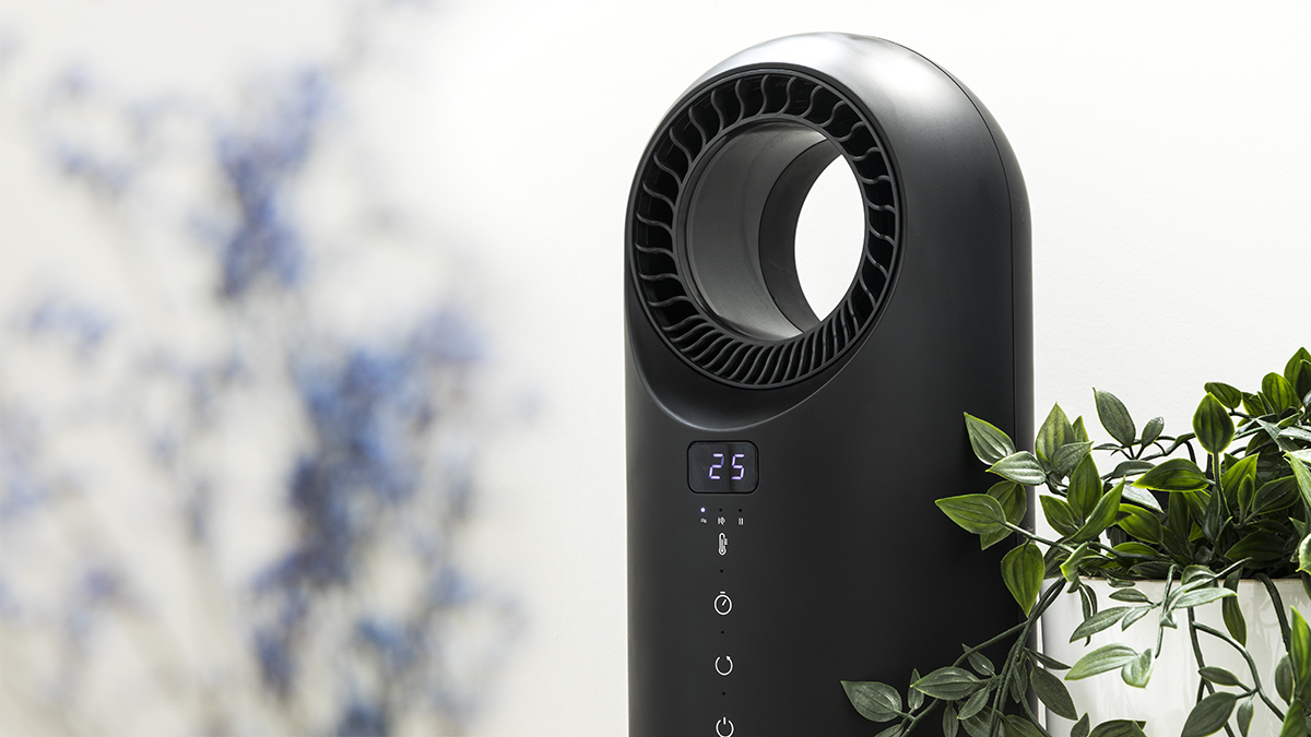 Purificador de aire negro con pantalla digital '25'. Planta verde en maceta blanca a la derecha. Flores azules borrosas al fondo.