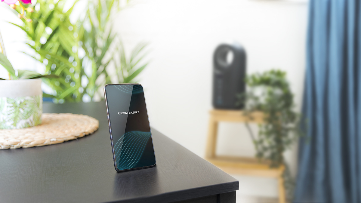 Un teléfono inteligente en una mesa negra muestra 'ENERGY SILENCE'. Plantas, una mesita con un purificador de aire y una cortina azul al fondo.
