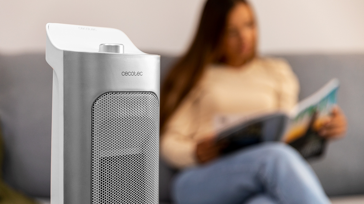 Un calefactor eléctrico plateado y blanco está enfocado. Una mujer lee una revista en un sofá en el fondo.