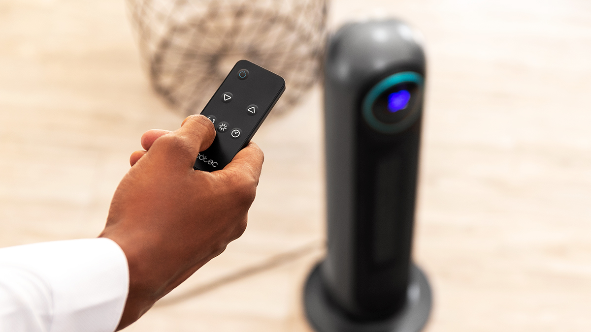 Una mano sostiene un mando a distancia, apuntando a un ventilador negro. Los botones del mando son visibles. El ventilador tiene una luz azul.