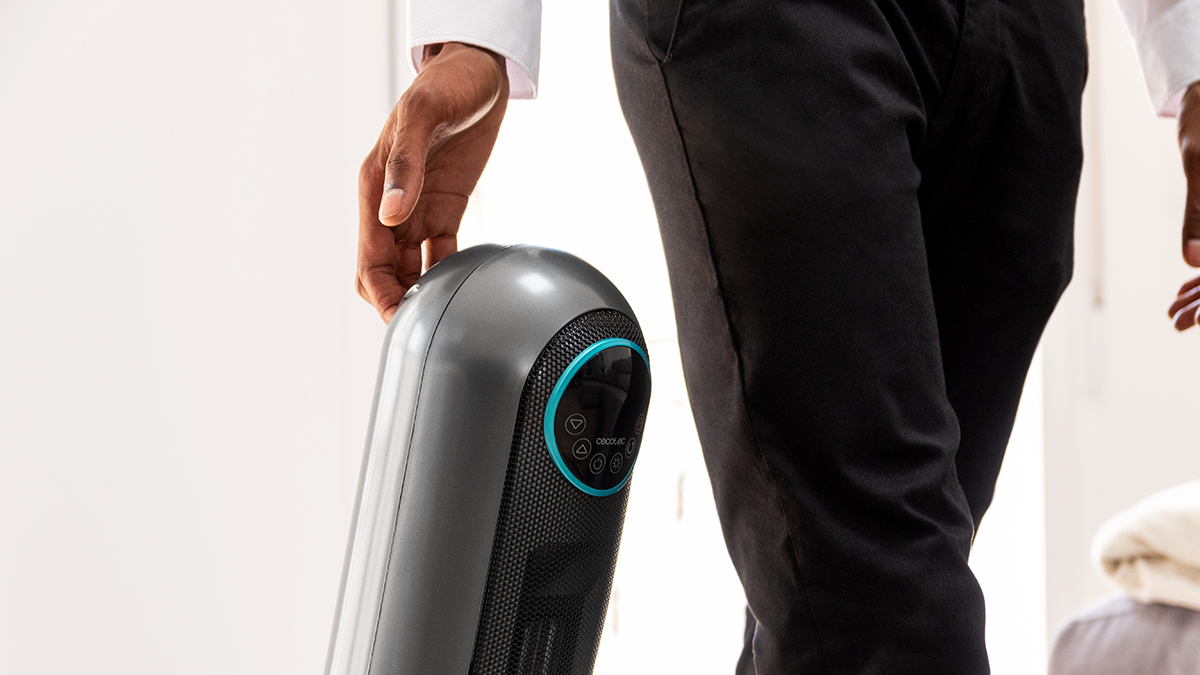 Una persona sostiene un calefactor eléctrico gris. El calefactor tiene una malla negra y un panel de control azul. La persona usa camisa blanca y pantalón negro.