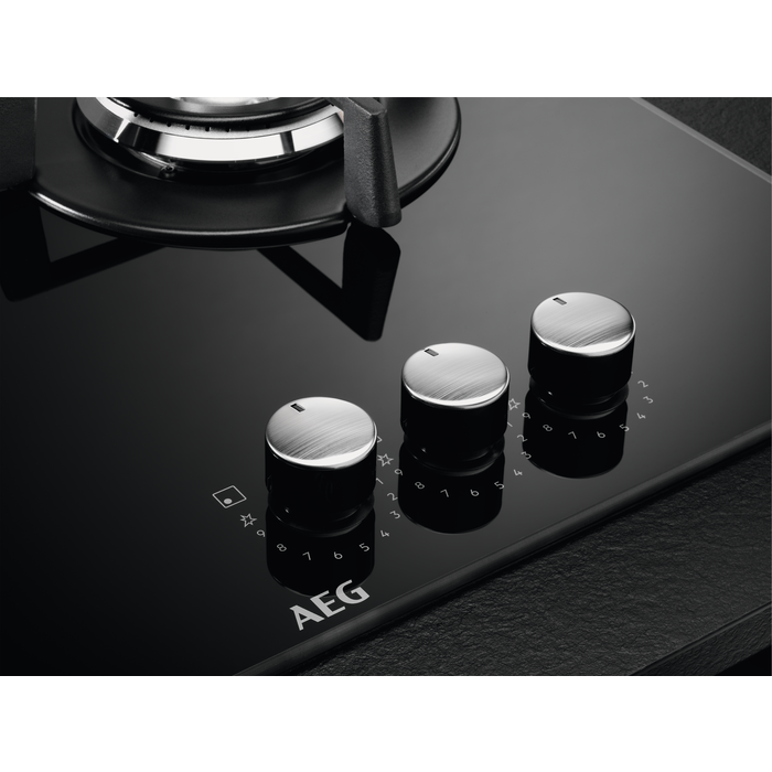 Primer plano de una placa de cocina de vidrio negro con tres perillas plateadas y la marca AEG.