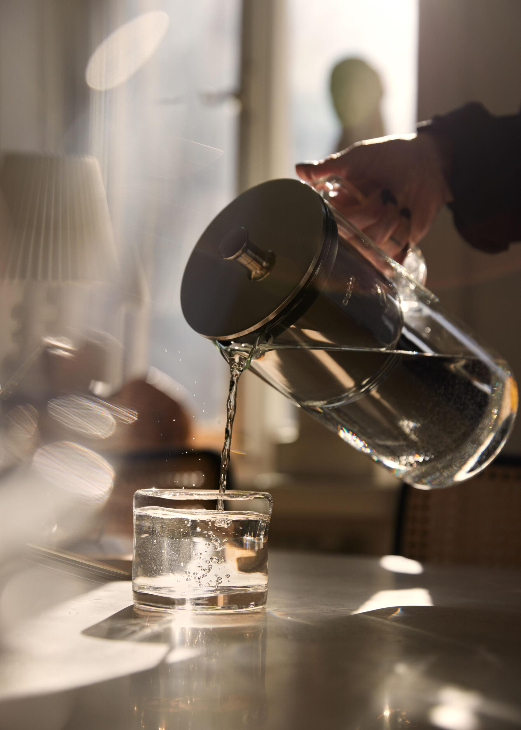 Eine Person gießt Wasser aus einer Glaskanne in ein Glas, Sonnenlicht scheint hindurch.