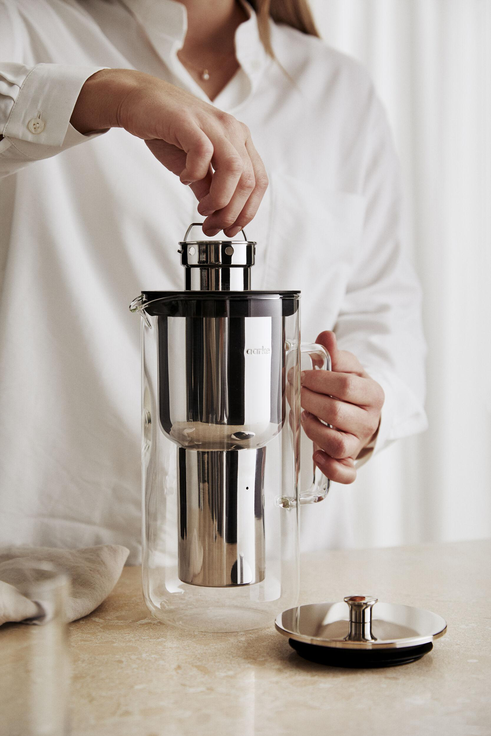 Hände einer Person, die einen Kaffeezubereiter aus Silber und Stahl halten.