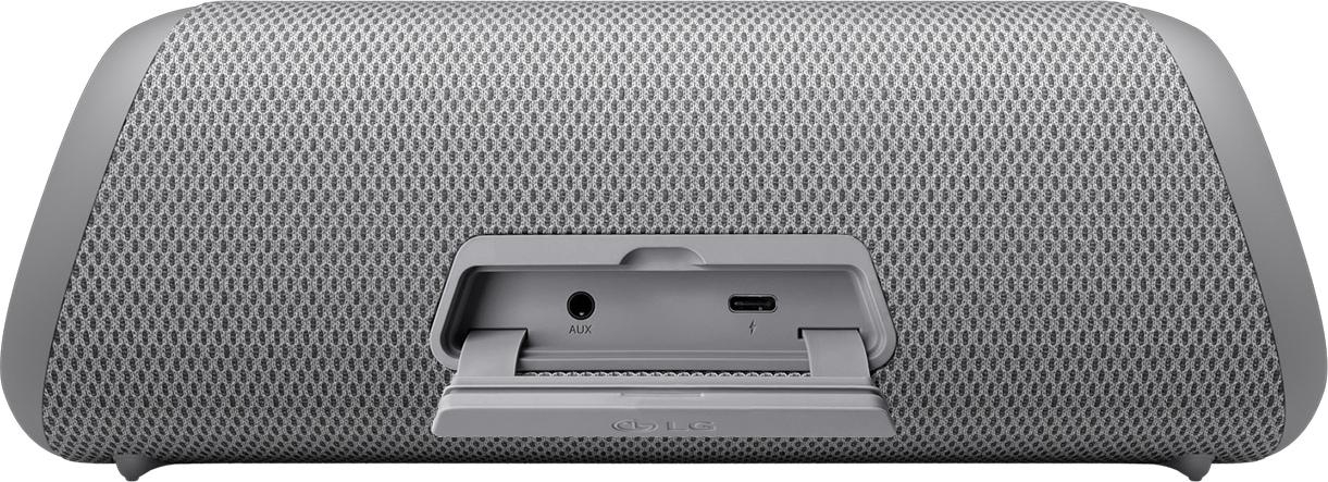 Grauer Lautsprecher mit Mesh-Front, kleiner Öffnung hinten mit Anschlüssen. Der Lautsprecher steht auf weißer Oberfläche.