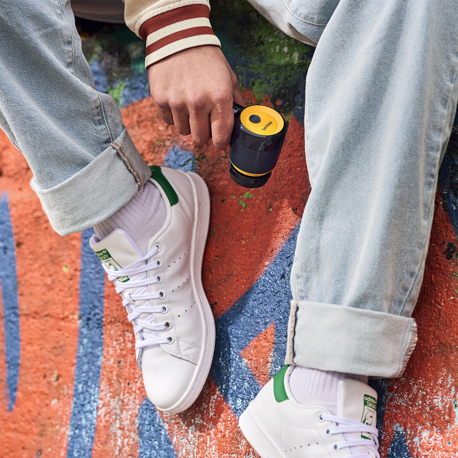 Una persona in jeans e scarpe da ginnastica bianche tiene in mano una fotocamera nera e gialla. Sfondo di graffiti.