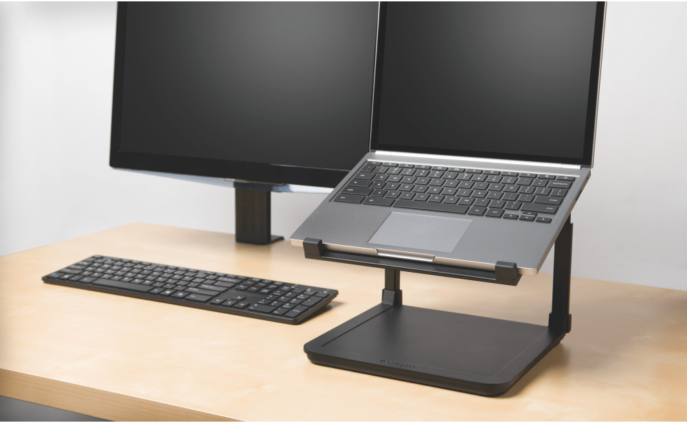 Laptop állványon, mögötte két monitorral és egy billentyűzettel. A laptop szögben áll, látszik a billentyűzet.