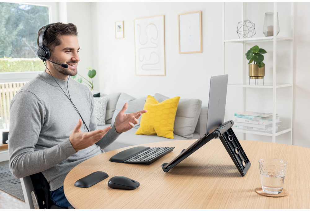 Férfi fejhallgatóval videohívásban, nyitott laptop, asztal billentyűzettel, egérrel és vízzel.