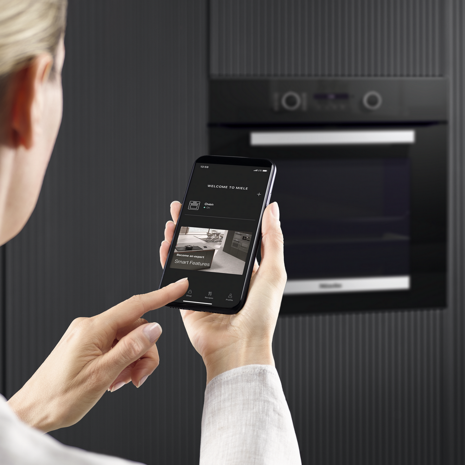 Woman uses smartphone in front of Miele oven, app shows 'Smart Features'.
