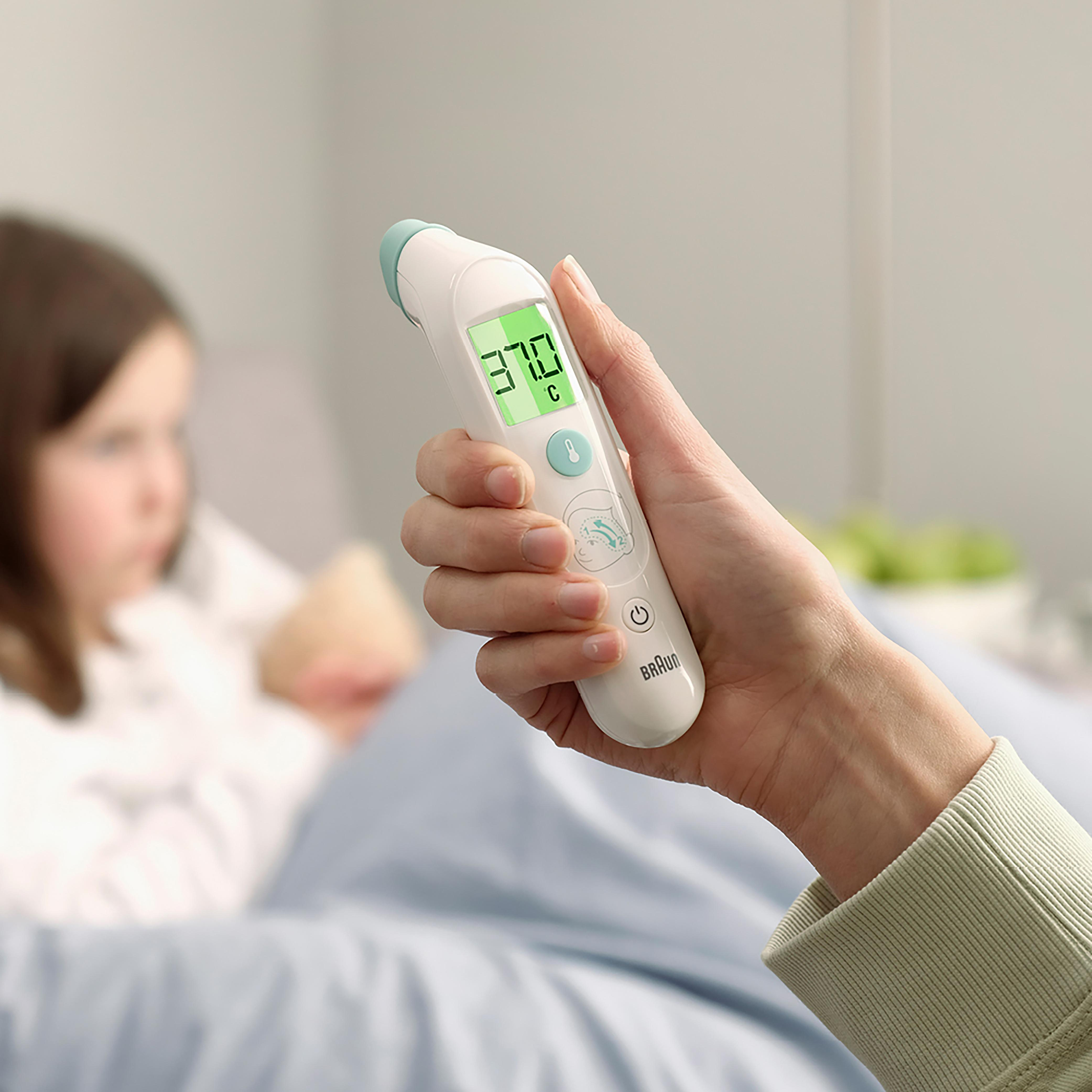 Eine Hand hält ein Braun-Thermometer, 37,0°C anzeigend. Ein Kind ist unscharf im Hintergrund.